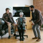 Two men assist a seated colleague with a virtual reality headset in a stylish, brick-walled office.